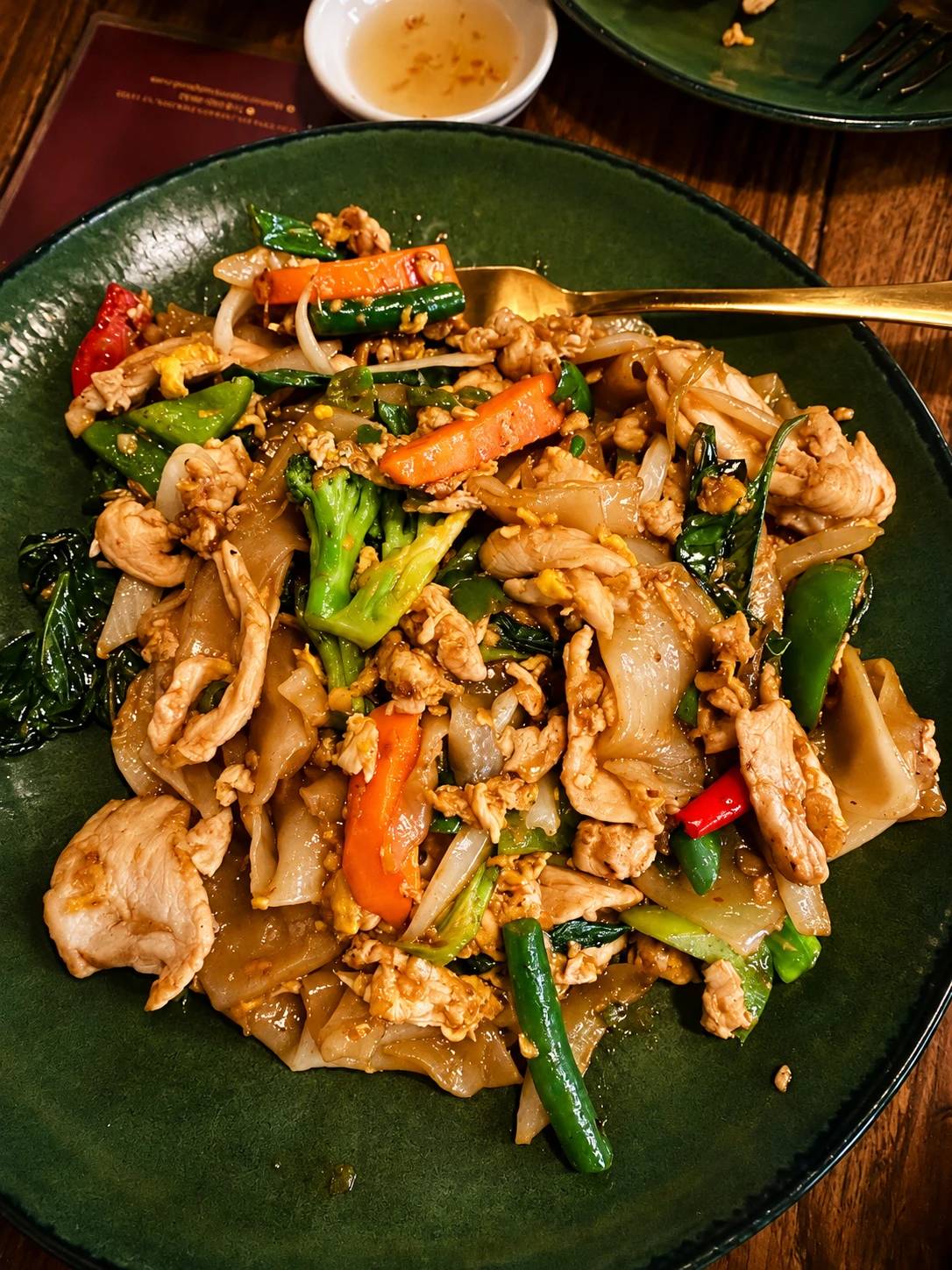 Broad rice noodles stir-fried with chicken, basil, broccoli, carrot and chili on a green plate.