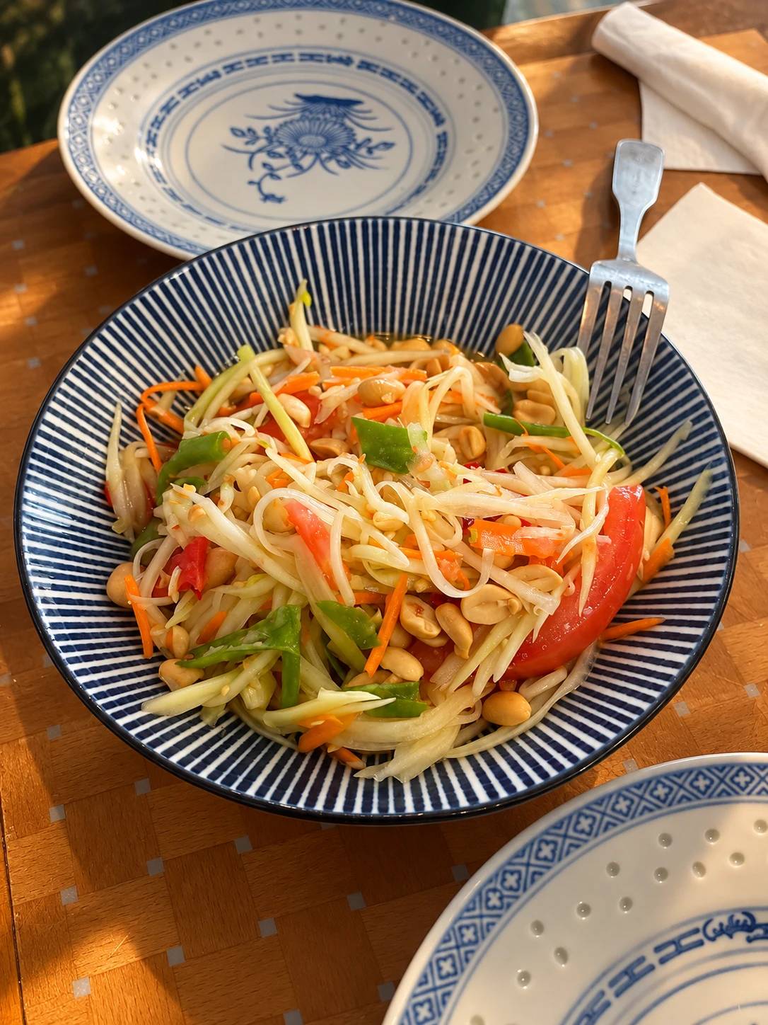 Green papaya salad with tomato, peanuts, carrot and green beans in a blue-striped bowl.