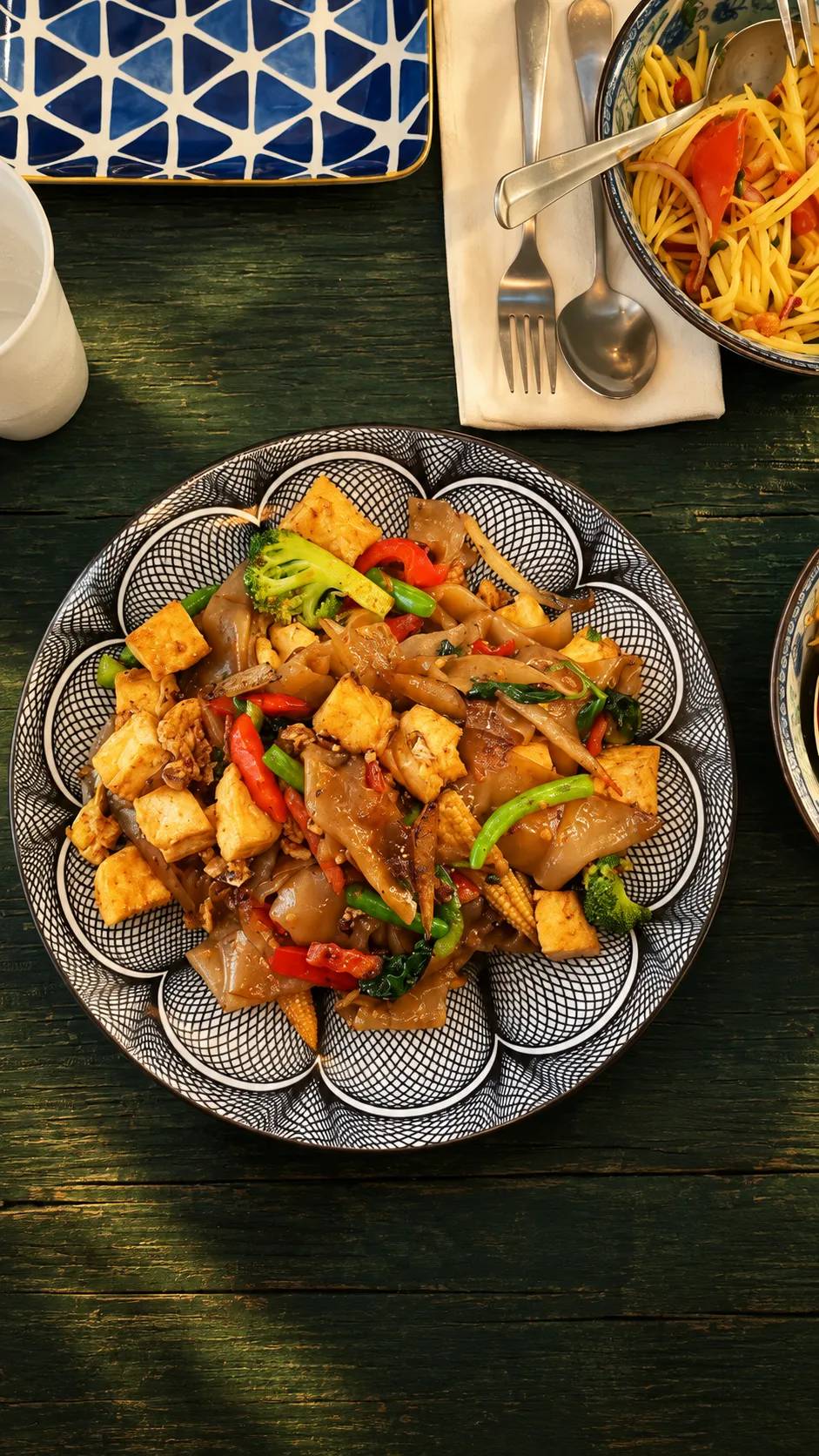 Broad rice noodles with fried tofu, broccoli, peppers and baby corn on a patterned plate.
